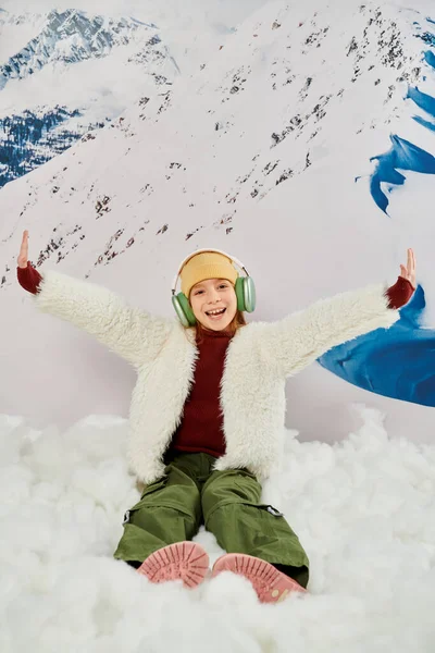 Vertical shot of girl in warm attire with headphones sitting on snow cheering with raised arms — Stock Photo