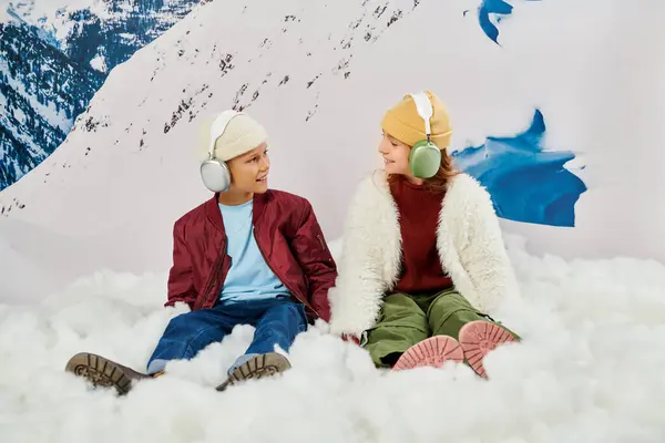 Lustige süße Jungen und Mädchen mit Headsets sitzen auf Schnee und lächeln sich fröhlich an, Mode — Stockfoto