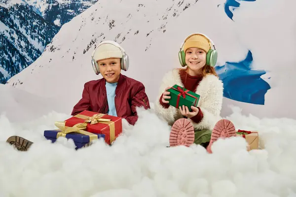 Cheerful smiley boy and girl in beanie hats sitting on snow with presents and headsets, fashion — Stock Photo