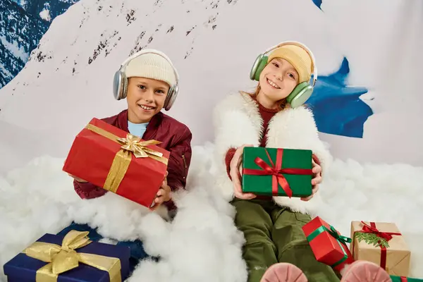 Allegri amici carini in abiti caldi con cuffie in possesso di regali e seduto sulla neve, moda — Foto stock