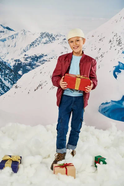 Vertikale Aufnahme eines fröhlichen kleinen Jungen in roter Jacke, der Geschenke hält und in die Kamera lächelt, Mode — Stockfoto