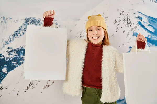 Gioiosa ragazza in cappello beanie e giacca invernale con in mano borse regalo e sorridente alla macchina fotografica, moda — Foto stock