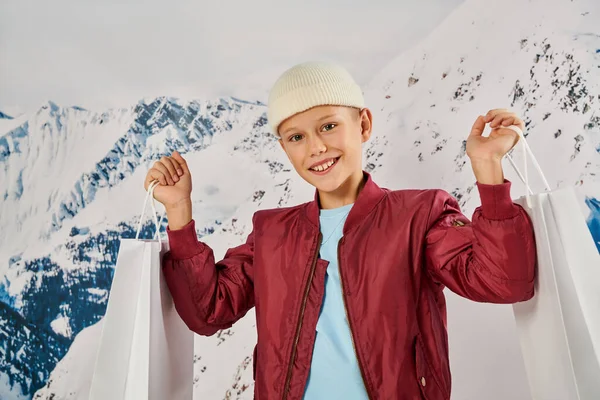 Kleiner Junge in stylischer Kleidung und Mütze mit Geschenktüten und lächelnd in die Kamera, Mode — Stockfoto