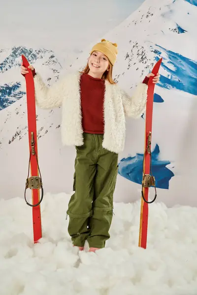 Vertical shot of joyous girl in stylish warm clothes holding skis with snowy mountain on backdrop — Stock Photo