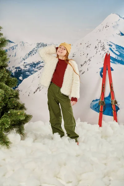 Vertikale Aufnahme eines fröhlichen Mädchens in stylischer Kleidung, das mit Skiern auf dem Rücken in die Kamera lächelt — Stockfoto