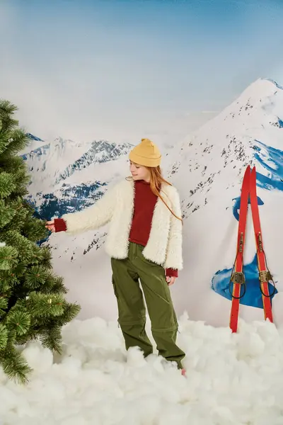 Plan vertical de petite fille mignonne touchant sapin avec skis et montagne enneigée sur toile de fond — Photo de stock