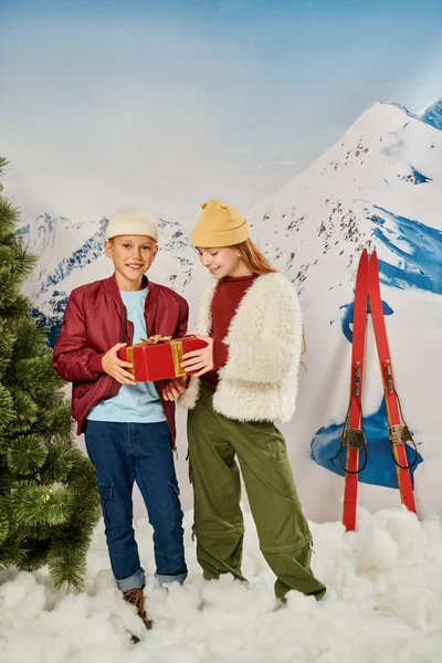 Vertical shot of preteen friends with present smiling happily with skis and mountain on backdrop — Stock Photo