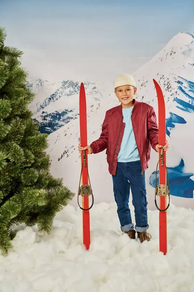 Tiro vertical de niño feliz en ropa elegante que sostiene esquís con montaña nevada en el telón de fondo - foto de stock