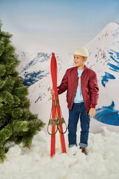 Tiro vertical de niño con estilo en traje cálido con esquís, sonriendo alegremente, concepto de moda - foto de stock