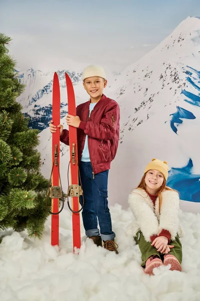 Colpo verticale di bambino in piedi e in possesso di sci accanto alla ragazza carina seduta sulla neve, moda — Foto stock