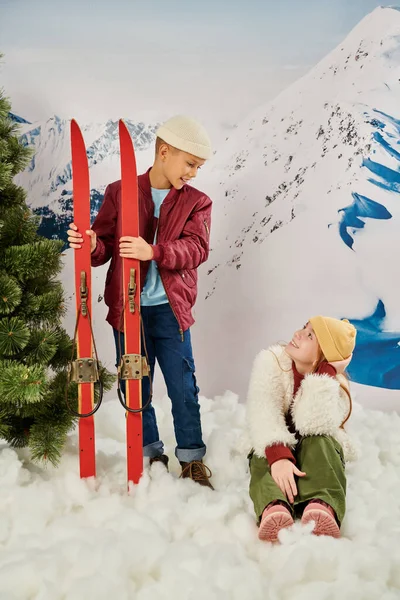 Tiro vertical de niña sentada en la nieve sonriendo a su amigo de pie sosteniendo esquís, moda - foto de stock