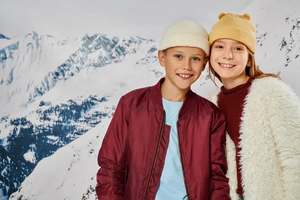 Preadolescent adorable children in winter attires smiling at camera with mountain backdrop, fashion — Stock Photo
