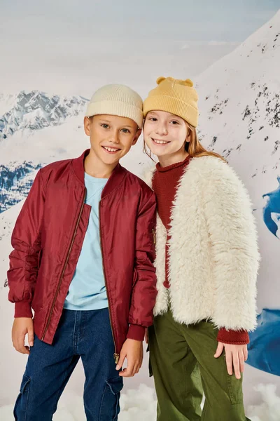 Alegre preadolescente chico y chica en ropa caliente de moda sonriendo a la cámara con telón de fondo de montaña - foto de stock