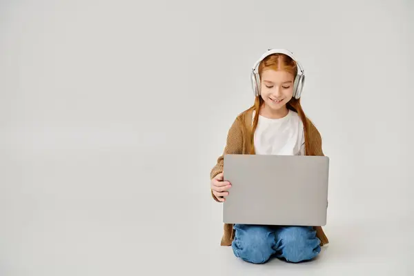 Joyful little girl in casual outfit with headset sitting on floor with laptop, fashion concept — Stock Photo
