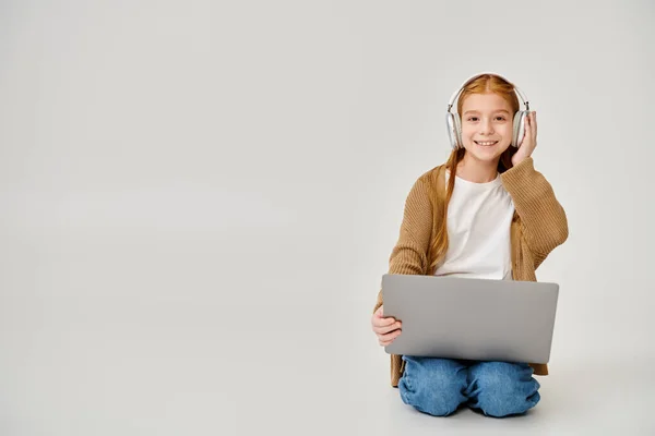 Happy little girl in casual attire with headset sitting on floor with laptop and smiling at camera — Stock Photo