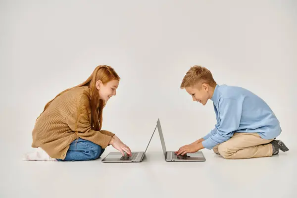 Cheerful preadolescent boy and girl in trendy winter outfits sitting on floor with laptops, fashion — Stock Photo