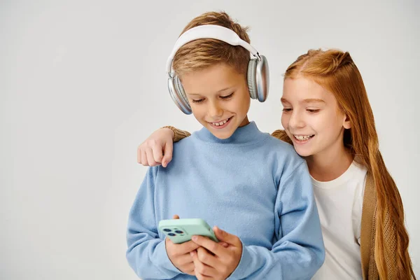 Red haired jolly preteen girl looking at phone of her cute peer with headset, fashion concept — Stock Photo