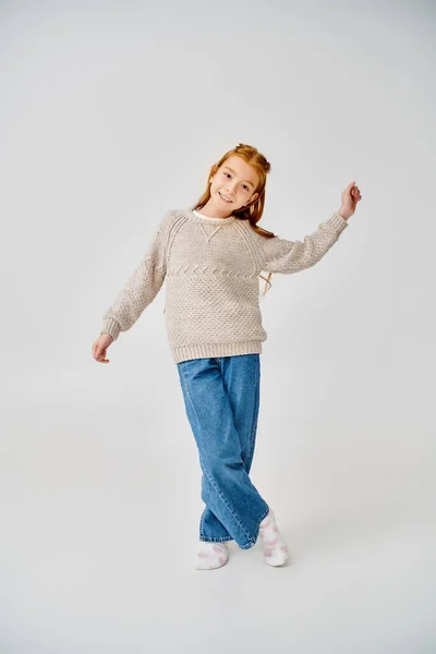 Веселая девочка-подросток в трикотажном свитере и джинсах позирует в движении на сером фоне, мода — стоковое фото