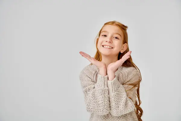 Bonita niña con el pelo rojo en suéter de punto casual mirando a la cámara con las manos debajo de la cara - foto de stock