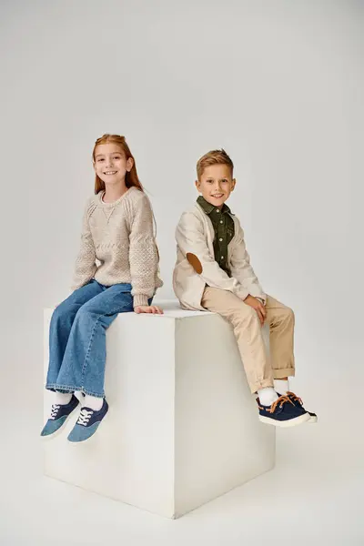 Vertical shot of two little children in casual trendy attires sitting on cube and smiling at camera — Stock Photo