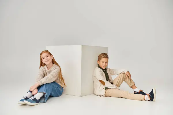 Happy preadolescent children sitting on floor next to white cube and smiling at camera, fashion — Stock Photo