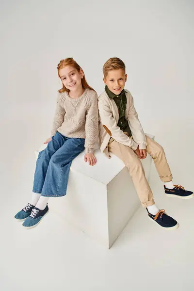 Cheerful preteen boy and girl in warm trendy clothes sitting on cube and smiling at camera, fashion — Stock Photo