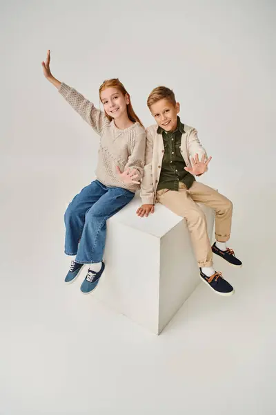 Plan vertical de joyeux petits amis en tenues d'hiver assis sur le cube blanc et agitant à la caméra — Photo de stock