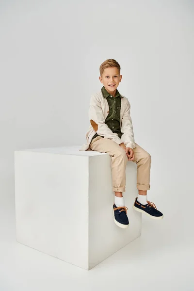 Vertical shot of joyous little boy in trendy cardigan sitting on white cube and smiling at camera — Stock Photo