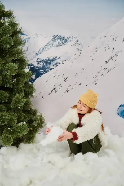 Весёлая рыжая маленькая девочка играет со снегом рядом с ёлкой на фоне горы — стоковое фото