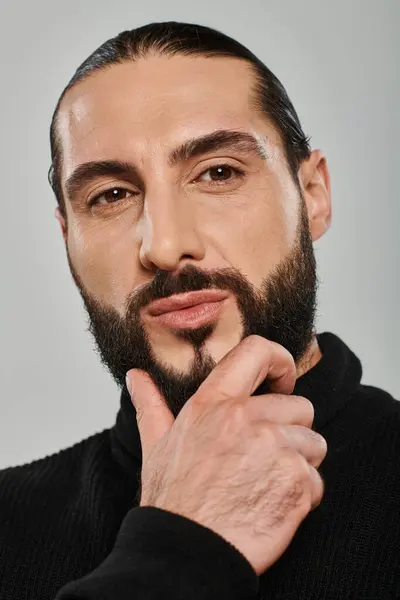 Retrato de hombre árabe guapo en cuello alto tocando la barba y posando sobre fondo gris - foto de stock