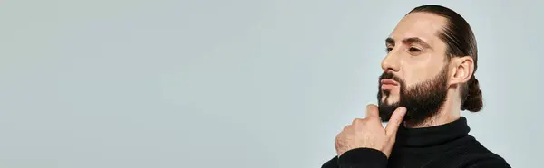 Portrait of handsome arabic man in turtleneck touching beard and posing on grey backdrop, banner — Stock Photo