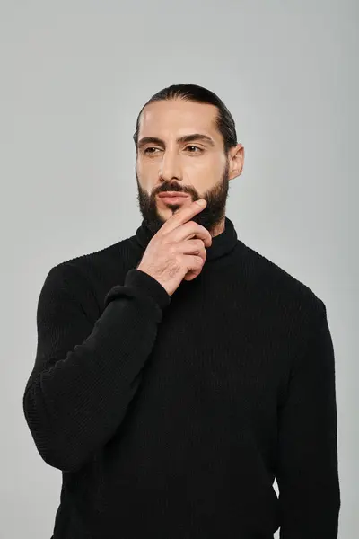 Retrato de hombre árabe guapo en cuello alto tocando la barba mientras pensaba en el fondo gris - foto de stock