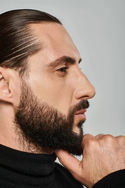 Profile of good looking arabic man in turtleneck touching beard while thinking on grey backdrop — Stock Photo