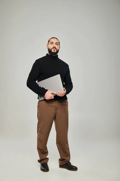 Uomo arabo bello e barbuto sorridente e in piedi con computer portatile su sfondo grigio, libero professionista — Foto stock