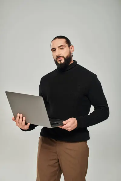 Bellissimo e barbuto uomo arabo in dolcevita nero in piedi con computer portatile su sfondo grigio — Foto stock