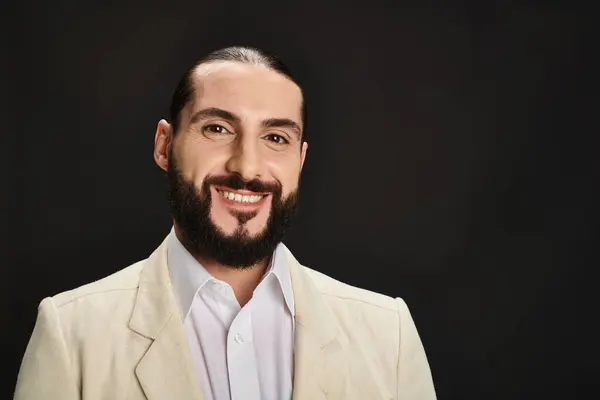 Hombre árabe alegre y barbudo en camisa blanca y chaqueta mirando a la cámara sobre fondo negro - foto de stock