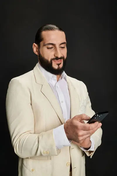 Fröhlicher und bärtiger arabischer Mann in weißem Hemd und Blazer mit Smartphone vor schwarzem Hintergrund — Stockfoto