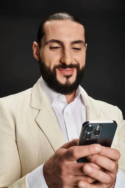 Allegro e barbuto uomo arabo in camicia bianca e blazer utilizzando smartphone su sfondo nero — Foto stock