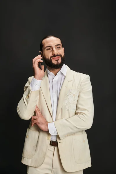 Hombre árabe alegre y barbudo en camisa blanca y chaqueta hablando en smartphone sobre fondo negro - foto de stock