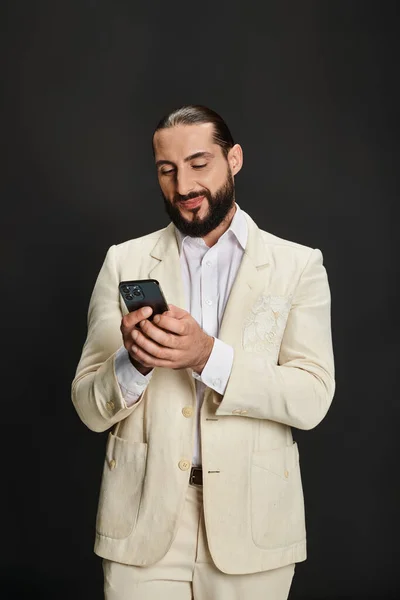 Fröhlicher und bärtiger arabischer Mann in weißem Hemd und Blazer, der vor schwarzem Hintergrund ein Smartphone textet — Stockfoto