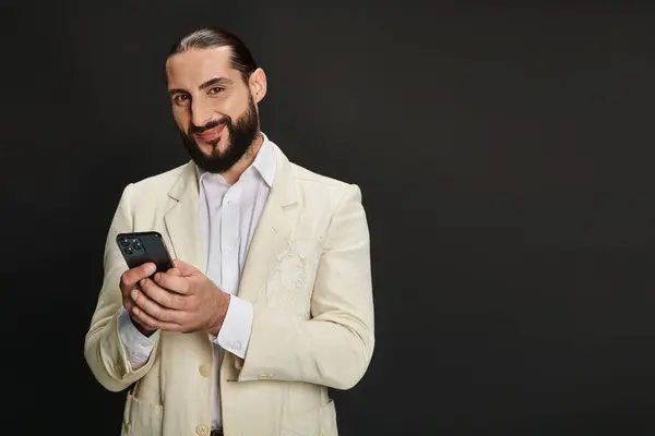 Hombre árabe alegre y barbudo en camisa blanca y blazer mensajes de texto smartphone sobre fondo negro - foto de stock