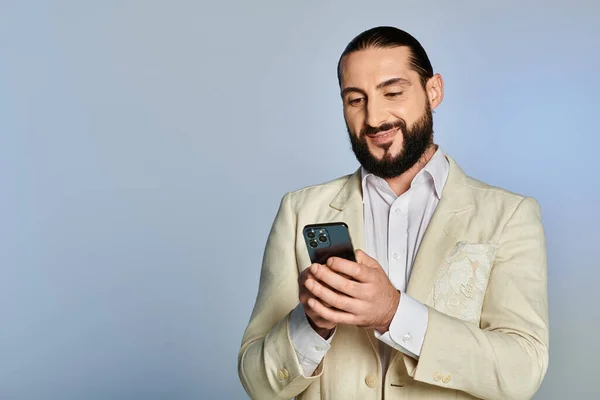 Glücklicher bärtiger arabischer Mann in weißer formeller Kleidung mit Smartphone auf grauem Hintergrund, elegante Kleidung — Stockfoto
