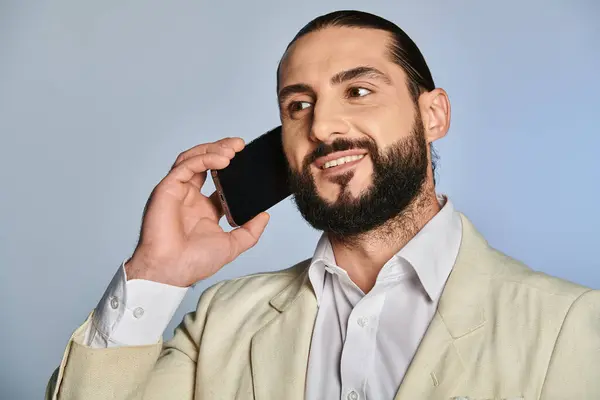 Hombre árabe barbudo feliz en ropa formal elegante hablando en el teléfono inteligente sobre fondo gris, llamada telefónica - foto de stock