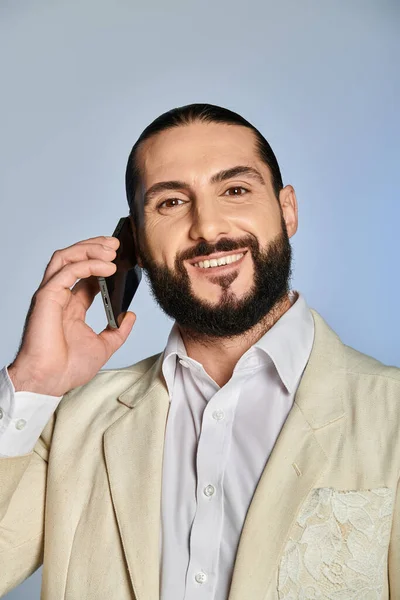 Hombre árabe barbudo feliz en ropa formal elegante hablando en el teléfono inteligente sobre fondo gris, llamada telefónica - foto de stock