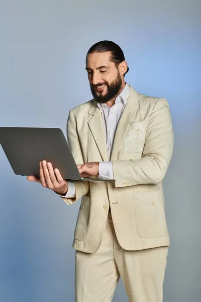 Felice uomo arabo barbuto in elegante abbigliamento formale utilizzando il computer portatile su sfondo grigio, era digitale — Foto stock