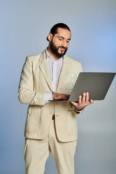 Felice uomo arabo barbuto in elegante abbigliamento formale utilizzando il computer portatile su sfondo grigio, era digitale — Foto stock
