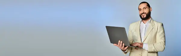 Felice uomo arabo barbuto in elegante abbigliamento formale utilizzando il computer portatile su sfondo grigio, banner dell'età digitale — Foto stock