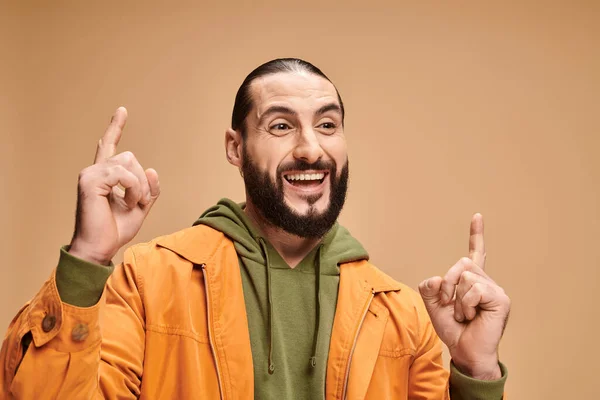 Portrait of positive and bearded arabic man in casual attire pointing up on beige background — Stock Photo