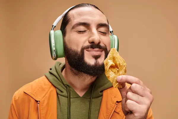 Happy arabic man in headphones smelling honey baklava on beige background, middle eastern dessert — Stock Photo