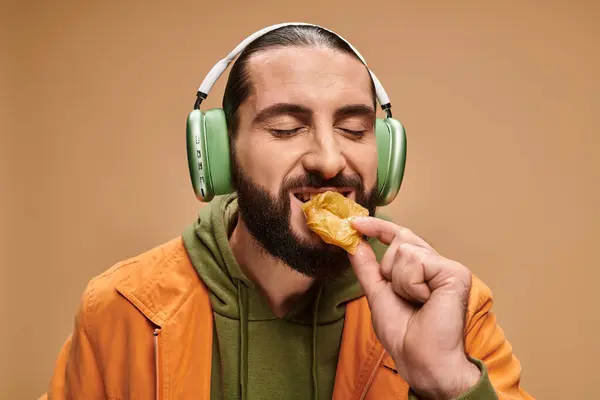 Cheerful man in headphones eating delicious honey baklava on beige backdrop, turkish delights — Stock Photo
